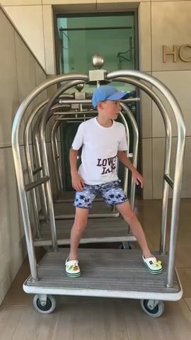 A young boy stands on a luggage cart. He is wearing a white t-shirt, patterned swim trunks, a blue baseball cap, and white clogs with green accents. He is looking to his right. The luggage cart is made of metal and has several wheels. Behind the boy, other luggage carts are visible. The background appears to be an indoor-outdoor area, possibly a hotel lobby, with a building entrance visible.