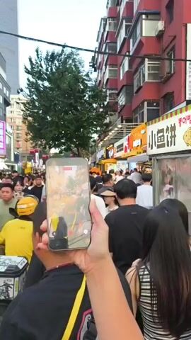 The video shows a crowded street scene, likely a market or a busy thoroughfare, with numerous people walking in both directions. The perspective is from behind a person holding up a smartphone to record the scene. The smartphone screen displays a live recording of the street, showing more people, stalls, and buildings. The people are dressed in casual clothing. The environment appears to be urban, with tall buildings on either side of the street and various signs and lights visible. The time of day appears to be late afternoon or early evening, as the light is soft and there are artificial lights illuminating the stalls. The overall atmosphere seems to be lively and bustling.