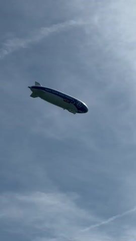 A blimp flies through the sky against a cloudy blue sky. The blimp is white with a blue stripe along its side. Text on the blimp reads "Next Generation Mobility. NOW." and a blue logo with "ZF" inside a circle is visible. A gondola is suspended beneath the blimp.