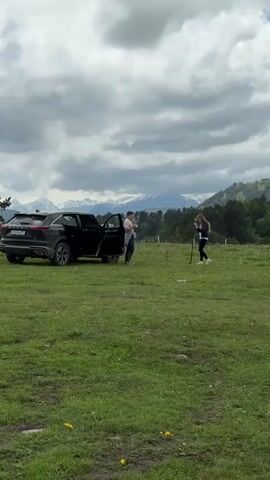 A black SUV is parked on a grassy hill with its passenger door open. A man stands by the open door, and a woman stands to his right, holding a tripod with a camera. The background features a forest and snow-capped mountains under a cloudy sky. The man appears to be talking to the woman, who is looking away from him. The woman then moves slightly to her right and lowers her camera.