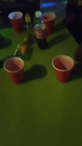 The video appears to be filmed at a bar or club in Barikin Sabon Gidan Kanat, Nigeria. The setting is dimly lit with a green table in the foreground and colorful disco lights in the background, suggesting it is nighttime.

Several red plastic cups are visible on the table, along with a bottle of Coca-Cola and what appears to be a bottle of alcohol. In the foreground, two individuals are partially visible, sitting at the table. One person is wearing a dark t-shirt with white text that appears to say "Valley" or something similar. This person is holding a bottle and looking towards the camera. In the background, more people are visible, and the atmosphere seems social and lively, with the disco lights creating a party-like ambiance.

The activity seems to be a casual gathering at a bar, possibly a party or social event, with drinks being consumed. There are no obvious signs of distress or negative emotions; the atmosphere appears relaxed and convivial. The time of day is clearly nighttime due to the artificial lighting. The location cues, like the people and the general vibe, are consistent with a bar or club in Nigeria.