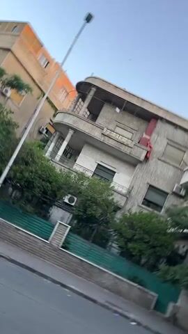 The video presents an urban street scene, likely in a residential area, under a clear blue sky on a bright day. The camera performs a slow panning motion, moving from right to left and then partially back.

In the foreground, a multi-story building dominates the view, characterized by its older, distinctive architectural style. This building features multiple balconies, including a prominent rounded balcony on the first floor supported by pillars, and an upper balcony from which a red fabric or banner hangs. Several windows, some fitted with blinds, are visible, along with an air conditioning unit on the ground level. A dense array of green trees and foliage occupies the space in front of this building, separated from a paved road by a low stone wall topped with a green metal fence.

In the background, a taller, multi-story building with a more modern appearance stands, its upper portion featuring an orange or brownish hue. A white street lamp pole extends upwards on the left side of the frame. A single bird can be observed flying briefly in the sky. No people are visible, and there is no discernible text on any of the objects or buildings. The overall scene suggests a quiet, established urban environment.