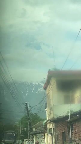 The video, taken from inside a vehicle, shows a view through a hazy windshield. Overhead power lines crisscross the sky. Snow-capped mountains are visible in the distance, partially obscured by clouds. Buildings and trees line the street, and a vehicle labeled "TONY" is visible in the foreground.