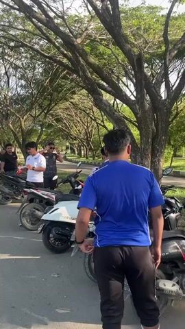 A group of men stand near a line of parked motorcycles under a canopy of large trees. The men are dressed casually in t-shirts and pants. Some are looking at their phones, while others appear to be talking. The motorcycles are parked on an asphalt surface, and there is grass and more trees in the background. The lighting suggests it is daytime, possibly late afternoon.