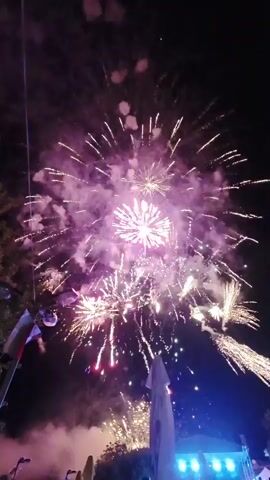 A fireworks display is captured at night. Several bursts of fireworks illuminate the dark sky with bright white and purple light. Smoke from the fireworks drifts downwards. In the foreground, there are silhouettes of people and trees. A white tent-like structure is visible to the right, and colorful lights are present in the background. The camera angle is looking upwards towards the sky.