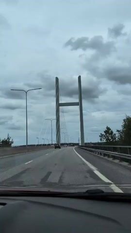 The video captures a drive over a bridge in Heinola, Finland, under a cloudy sky. The initial segment shows the car approaching a prominent cable-stayed bridge with two tall, grey pylons and numerous supporting cables. Other vehicles are also on the multi-lane road. The camera then shifts to a side view from inside the car, looking out the passenger window. A red car's side mirror is visible, reflecting a portion of the vehicle. Beyond the bridge railing, a calm body of water, likely a lake or river, stretches towards a forested shoreline in the distance. The sky remains overcast throughout the video.