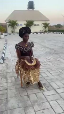 A young, slim Black woman sits in a wooden chair on a paved patio, wearing traditional African dress. She has a large, black head covering, and her dress is dark brown on top, with a leafy pattern over the shoulders. She wears a gold pendant necklace and a gold ring. The lower part of her dress is gold and glittering with a brown feathered wrap over her knees. She holds a matching feathered fan. She wears gold shoes. Behind her is a one-story building with a light-colored roof and a water tank on top. There are palm trees and other plants in the background. The sky is pale. The camera is stationary.