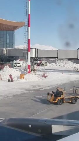The video shows an airport scene in winter from the perspective of an airplane window. The ground is covered in snow. In the foreground is part of the airplane wing. A red and white striped pole stands near the airport terminal. Behind it are various airport support vehicles, including a white van and a yellow utility vehicle with a flatbed. The airport terminal building is connected to the gate by a jet bridge. Mountain peaks are visible in the distance. There is no evidence of people interacting.