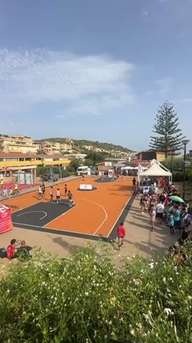 The video shows an outdoor basketball court on a sunny day. The court is orange with black lines. Several people are playing basketball, and a crowd is watching from the side. There are buildings in the background, and some trees. A white tent is set up near the court. The sky is blue with some clouds.