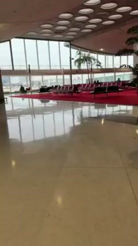 The video shows an autonomous trash receptacle robot moving across a bright tile floor, likely in an airport. The robot is black with blue lights underneath, and has a display screen with information on it, and a receptacle for trash. It is moving toward a large waiting area with rows of chairs on a red carpet. Large windows overlook an airfield. Several people are visible in the waiting area. The ceiling is high with a pattern of circular lights.