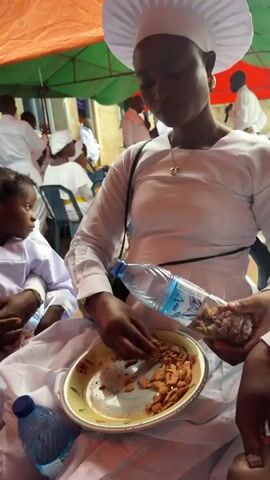 The video shows a close-up of a woman in white religious attire and a white hat. She is seated and holding a partially filled plastic water bottle and a plate of food. She is using her fingers to pick up pieces of food from the plate and place them into the bottle. In the background, other people in similar white attire are visible, some seated, some standing. There is an orange and green tarp overhead, suggesting an outdoor or semi-outdoor setting. The overall scene appears to be a gathering or event related to a religious ceremony.
