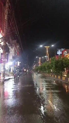The video captures a street at night with wet pavement, suggesting it has recently rained. The street is lined with buildings on the left and trees on the right, with decorative streetlights illuminating the scene. Several motorcycles are parked along the sidewalk on the left. There are no visible people engaged in significant actions or interactions. The environment appears to be an urban street.
