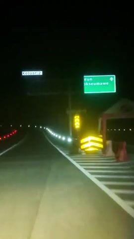 The video shows a first-person perspective from inside a moving vehicle on a highway at night. The road is well-lit by streetlights and the vehicle's headlights. On the right side of the road, there's a green sign with white text that reads "eun Lhokseumawe" and an arrow pointing to the right, indicating an exit. Below this sign, there's a yellow directional sign with black arrows. Further down the road, on the right, there are yellow and black striped barriers. On the left side of the road, another sign can be partially seen with the text "Keluar 2," which translates to "Exit 2." The road has white solid lines on both sides and a yellow solid line separating the two lanes. A row of bright white lights lines the right shoulder of the road. No other vehicles or people are visible in the immediate vicinity.