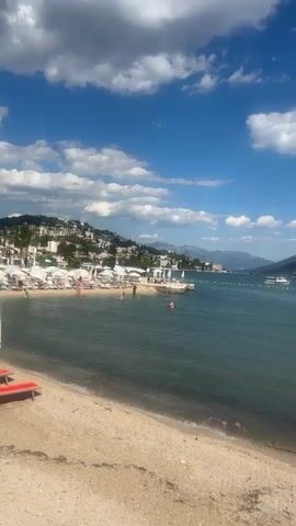 This is a vertical, POV video filmed at a beach. The video begins with a wide shot of a bay with a clear blue sky and scattered white clouds. The water is calm and appears to be a moderate blue-green color. A sandy beach is in the foreground, with some lounge chairs and umbrellas. In the mid-ground, there are several people swimming in the water, some on inflatable devices, and a few boats are anchored in the bay. Along the shoreline, there is a resort or town with white buildings nestled into a green, hilly landscape that stretches into the distance. The video pans slowly to the right, showing more of the bay, the distant hills, and the beach. In the foreground, two orange and red striped lounge chairs with a white and light blue umbrella are prominently displayed. A person is sitting in one of the chairs, facing away from the camera. The video continues to pan right, revealing more of the beach with additional lounge chairs and umbrellas, and a rocky embankment to the far right. The overall scene depicts a sunny day at a seaside resort, with people enjoying recreational activities.