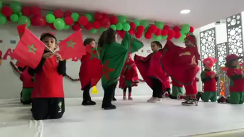 A group of approximately ten young children perform on a stage decorated with red and green balloons. The children are dressed in red and green outfits and are waving small Moroccan flags. Some of the children are kneeling while others are standing and dancing. The stage has the words "IETS DE CLASSES" written on its front. The background features a wall with a decorative pattern and an array of international flags. The room is brightly lit.