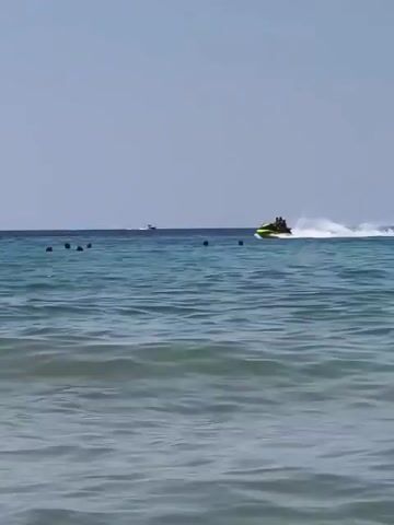The video shows a body of water with a clear blue sky. In the distance, a yellow jet ski is being ridden by at least two people, creating a white spray of water as it moves. Closer to the viewer, several people are swimming in the water. The water is a deep blue with some waves visible in the foreground. The overall scene depicts a recreational day at the beach.
