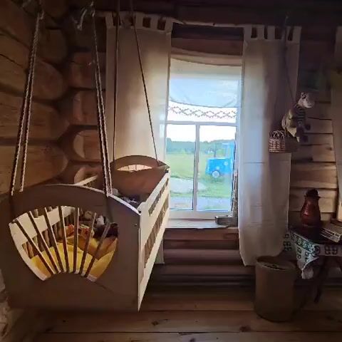 This video shows a wooden crib hanging from ropes in a rustic, log-cabin-style room. The crib is filled with yellow bedding and what appear to be stuffed animals or toys. A large window with white curtains is visible behind the crib, offering a view of a green field with a blue object in the distance. To the right of the window, a small table holds a red lantern and other items. The floor and walls are made of wood. The scene is dimly lit, with light coming from the window. There are no people visible in the video. The crib appears to be gently swaying, suggesting it is in motion.
