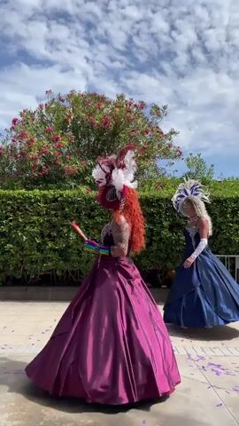 The video shows two individuals in elaborate costumes. The person in the foreground wears a voluminous dark pink gown and a large, feathered headdress in red and white. They are holding a rainbow-colored fan in their left hand and appear to be looking away from the camera. The second individual, further back and to the right, is wearing a dark blue gown and a white and blue feathered headdress. They are looking towards the first individual and holding a red rose. Both individuals appear to be standing in an outdoor setting with a manicured hedge and a flowering tree in the background, under a cloudy blue sky. Confetti is scattered on the ground.