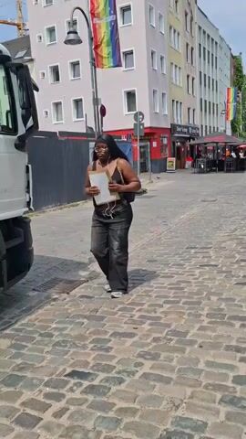 A young Black woman wearing glasses, a black top, and baggy grey jeans walks past a white truck with red lettering on its side. She is carrying a light-colored box and a black purse. The truck appears to be a delivery vehicle, with the partial word "GOURMET" visible in large red letters on its side. The woman is walking on a cobblestone street, and in the background, there is a building with a rainbow flag displayed on a pole. The scene suggests an urban setting, possibly during the day, with fair weather. The woman's expression is neutral as she walks. The overall impression is that of an everyday scene in a city.