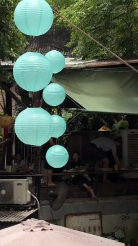 The video displays an outdoor setting, possibly a cafe or restaurant, with several people present. Several light blue paper lanterns are hung in the foreground, obscuring parts of the background. In the mid-ground, there are tables and people sitting at them. One person is standing at a table, appearing to interact with food or objects on the table. Another person is seated at a table, looking towards the foreground. An air conditioning unit is visible on the left side of the frame. The environment is lush with green foliage in the background. The overall impression is a casual, outdoor gathering space.