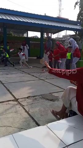 This video shows an outdoor activity with several children participating in a race or game. The scene appears to be a paved area with a covered structure in the background. Several adults are present, some of whom seem to be supervising or cheering on the children. The children are running, and some are holding small flags or objects. The weather appears to be sunny. The activity involves a series of buckets placed on the ground.