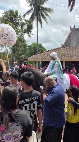 A man in white Islamic attire, including a white turban and a green sash, rides a dark horse adorned with red and white blankets. He holds a large, round white flower decoration above his head, which is attached to a pole. The horse is surrounded by a crowd of people, some of whom are looking at the rider. To the left of the horse, another person in Islamic attire, wearing a hijab, is also visible. The scene appears to be outdoors, with a blue sky and clouds visible overhead, along with palm trees and a telecommunications tower in the background. The ground is dusty, suggesting an unpaved area. Some people in the crowd are holding their phones up, possibly to record the event. The overall atmosphere suggests a procession or a cultural event.