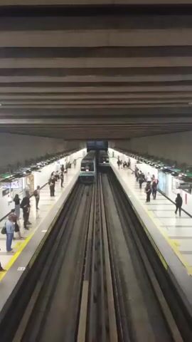 The video shows a subway station platform with two sets of tracks and people waiting. A train, predominantly blue and white, arrives on one of the tracks. The train has the number "053" displayed on the front. People are standing on both sides of the platform. The station is underground, with a low, horizontally striped ceiling. The platform has yellow safety lines.