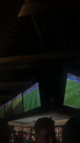 The video shows a dimly lit room with multiple large flat-screen televisions mounted on the ceiling, all displaying a soccer game. The game appears to be in progress, with players visible on the green field. The lighting is low, creating a dark ambiance, with the screens being the primary source of illumination. In the foreground, the back of a person's head and shoulders is visible, suggesting that the viewer is seated or standing below the televisions. The overall environment seems to be an indoor venue, possibly a sports bar or a similar establishment where people gather to watch sports.