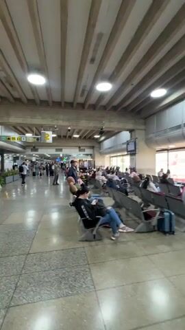 The video shows an airport waiting area with people sitting on chairs while others walk by. The waiting area has a row of chairs on either side of a walkway. Many people are using phones, reading, or sleeping. A sign overhead indicates gate 2. The architecture of the building features a high ceiling with exposed concrete beams. There are shops and restaurants visible in the background.