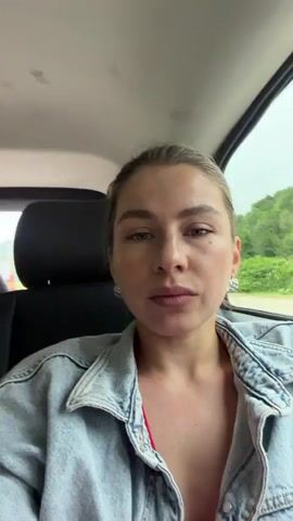This is a close-up, selfie-style video of a woman sitting in the passenger seat of a car, likely in motion. She has her hair pulled back and is wearing a denim jacket over a red top. Her eyes are looking directly at the camera with a serious expression. Her right hand is resting on her chin, and her fingers are extended. The background is blurred, showing greenery and trees outside the car window, suggesting a rural or natural setting. The sky appears overcast, indicating cloudy weather. The overall mood is contemplative or perhaps a bit somber. No visible text is present in the video.