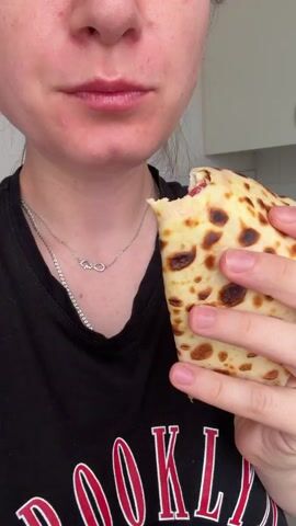 The video shows a close-up of a person eating a rolled-up crepe or thin pancake. The person is wearing a black t-shirt with red lettering that says "BROOKLYN". They are holding the crepe with their right hand and taking a bite. The crepe appears to be filled with a mixture of ingredients, including what looks like diced tomatoes and possibly cheese. The background is blurred but suggests an indoor setting, possibly a kitchen, with white cabinets visible. The lighting is bright.