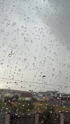 A vertical video shows a windowpane covered in raindrops. The sky is overcast and grey. In the background, out of focus, are several houses with visible roofs and a dark fence. The overall impression is of a rainy day.