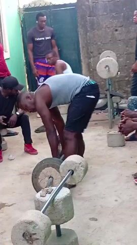 The video shows a man lifting a barbell with concrete weights in what appears to be a backyard or courtyard. Around him are several other men, some sitting and watching, others standing. The man lifting the weights bends over to grip the bar, then lifts it off the ground. He struggles briefly before setting it back down.  The men are of varying heights and builds, dressed in casual clothing. The surroundings include a green painted building with a white window frame, a concrete wall, and scattered debris on the ground. The atmosphere seems relaxed and supportive.