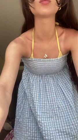 A young woman with long brown hair is modeling a light blue and white checkered dress in a hotel room. The dress has a smocked bodice and a halter top with yellow straps that tie at the back, leaving the back partially exposed. She has two small pink flower clips in her hair and is wearing a gold necklace. The room has a beige wall, a patterned bedspread, and a bedside table with various items on it. The woman spins and poses for the camera.