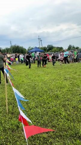 A group of children are participating in a race outdoors on a grassy field. They are running towards the right side of the frame, and several spectators are watching them from the background. There are cars and tents set up behind the spectators. The children are wearing casual clothing, and some of them are holding sticks. The race appears to be part of an outdoor event or festival, with colorful flags decorating the area. The sky is cloudy.