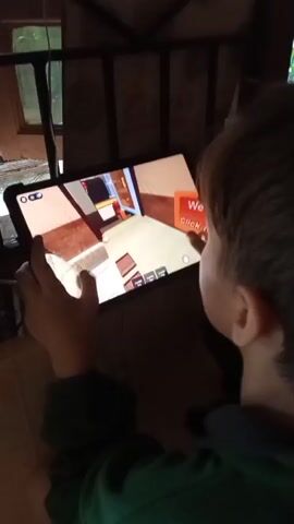 This is a close-up, handheld video of a child playing a game on a tablet. The child is seen from behind, wearing a green hooded sweatshirt. Their hands are on the tablet, fingers actively interacting with the screen. The game displayed on the tablet appears to be a 3D adventure or role-playing game with blocky character models and colorful environments. Text visible on the screen includes "ROBLOX" in the upper left corner and some game-specific text and icons. The setting seems to be indoors, possibly a room with natural light coming from a window visible in the background. The overall mood is one of focused engagement by the child in their game. The context provided indicates the location is Paliem, India, suggesting a tropical or subtropical environment, though this is not explicitly evident in the image itself beyond the natural light.