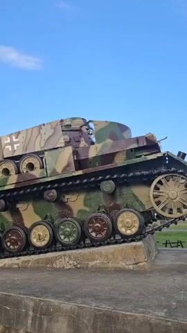 A camouflaged tank is positioned at an angle on a stone base. The tank has a German cross insignia on its turret. Another green tank is positioned to the left of the camouflaged tank, also on a stone base. A man in a red shirt and black shorts is standing on top of the turret of the green tank. He is looking away from the camera. The sky is blue with some white clouds.