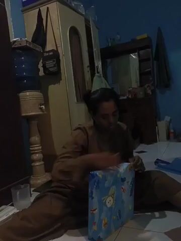 The video opens with a shot of a woman sitting on the floor, wearing a brown long-sleeved shirt and pants, handling a blue and white container. A water dispenser on a wooden stand is visible to her left, and a dark wooden cabinet with a reflective surface is in the background. The camera then pans quickly and settles on a man sitting on a blue mattress or couch, wearing a dark t-shirt and dark pants. He is looking down at a white smartphone. His hair is dark and disheveled. The same wooden cabinet is visible behind him against a light blue wall.
