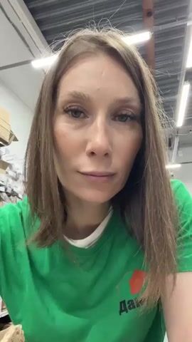 A young woman with long brown hair and fair skin is filming herself. She is wearing a bright green t-shirt with a logo that appears to be an orange diamond shape with text underneath. Her nails are painted white. Her left hand is in the foreground, and she is holding up two fingers in a peace sign. She then moves her fingers together as if to indicate a small size. The background appears to be an indoor setting, possibly a warehouse or storage area, with shelves and boxes visible. The lighting is bright, coming from overhead fluorescent lights.