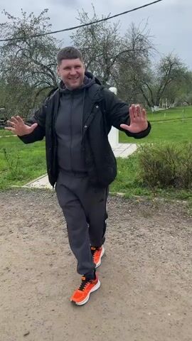 A man wearing a black jacket, gray tracksuit, and orange sneakers is dancing on a gravel path. He is wearing a backpack and facing towards the camera with a smile on his face. The background is a grassy field with trees and a white gazebo in the distance. The sky is overcast.
