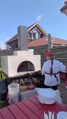 A woman wearing a red cap, white shirt, and apron, with her hair in a braid, is standing in front of a large wood-fired oven. She is wearing gloves and appears to be preparing food. There are bowls of ingredients and a pan on a table in front of the oven. Behind the oven, a rustic wooden building with a red roof is visible. The scene is outdoors with a clear blue sky.