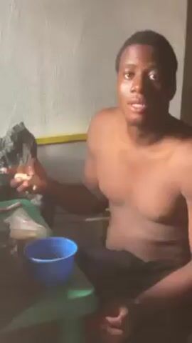 A shirtless Black man sits at a green plastic table. A blue plastic bowl is on the table, with a black plastic bag next to it, and a clear plastic bag with something inside. The man looks at the bowl and seems to be holding something in his hand. The background consists of white walls, with a glimpse of what appears to be a patterned curtain. The lighting in the room appears dim.