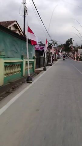 The video shows a view from a moving vehicle on a paved road. The road is lined with Indonesian flags. On the left side of the road, there are houses and buildings, some with green fences. On the right side, the road curves and continues into the distance with more flags and buildings. A few motorcycles are visible on the road. The sky is cloudy.