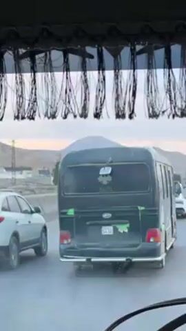 The view is from inside the front of a vehicle, looking through the windshield, at a dark green bus with a four-leaf clover decal on the back. The bus is in the lane in front of the camera car, with a white SUV to its left. Both vehicles are moving in a line of traffic on a multi-lane highway. Behind the cars, in the distance, is a brown, rocky mountain. Above the windshield is a decorative fringe.