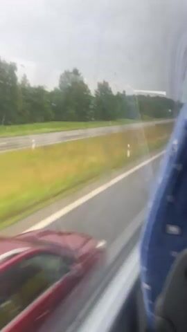 The video shows a view from inside a vehicle, likely a bus, looking out a window. The window has several scratches and water droplets on it, indicating it is raining or has recently rained. Outside, a red station wagon is driving on a wet highway. The highway is bordered by green trees and fields. The interior of the vehicle is visible in the foreground, showing a blue seat with a patterned fabric. The perspective suggests the viewer's vehicle is also moving, given the motion blur of the car and the landscape. The weather appears overcast.