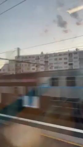 The video shows a view from a train window.  A train pulls into a station, blurring as it slows. Outside, other train cars are visible, with a multi-story building in the background under a cloudy sky. The train windows are reflective.