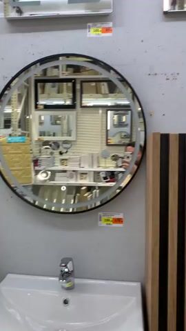 The video pans across a store display featuring bathroom sinks and mirrors. In the first shot, a man is visible in a round mirror, taking a selfie. He has dark hair, a beard, and a tattoo on his arm. He is wearing a light-colored t-shirt. The mirror has an LED light strip around its perimeter. The background of the mirror's reflection shows shelves stocked with various items, including smaller mirrors. The next shot shifts focus to a rectangular mirror above a white sink. This mirror also has an LED light strip. The reflection in this mirror shows the same store environment with shelves and other mirrors. The final shot moves to the right, showing a wooden cabinet and a close-up of a blue product tag hanging from a shelf. The overall environment appears to be a retail store, likely a home improvement or bathroom supply store, with bright overhead lighting and concrete walls.