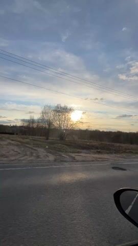 The video shows a view from inside a moving vehicle, likely a car. The camera is pointed out the side window, capturing a landscape passing by. A two-lane road with a white dividing line is visible in the foreground. Beyond the road is a strip of land with some vegetation, including trees without leaves. In the background, a line of trees is silhouetted against the setting sun, which is partially obscured by clouds in the sky. Power lines run parallel to the road above. The overall scene has a serene and peaceful quality, typical of a sunset drive.