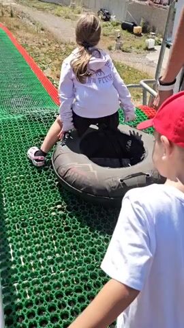 A young boy in a white t-shirt and red baseball cap stands on a green, mesh-like surface, facing away from the camera. To his right, a girl in a white hoodie is seated on a black inner tube, looking slightly to her left. A person's arm, wearing a grey sleeve and white sneakers, is visible near the girl, possibly assisting her. The background shows a long, sloped area covered with green and red material, a parking lot with cars, and a mountainous, tree-covered landscape. The overall scene suggests an outdoor recreational area.