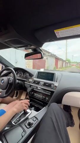 This is a first-person perspective video taken from inside a car, likely a BMW, as evidenced by the steering wheel logo. The driver, a person wearing blue shorts, is seen operating the vehicle. Their hands are visible on the steering wheel and the gear shifter. Another person, wearing black pants, is in the passenger seat, with their legs visible in the foreground.

The car is driving through a street lined with garage doors. These garages appear to be made of brick and painted in various colors, mostly red and white. The road surface is paved. The sky is overcast, suggesting a cloudy day, and the overall lighting is diffused. The time of day appears to be daytime.

A warning sticker with a triangle and exclamation mark is visible on the sun visor, written in Russian. The text on the sticker is mostly illegible due to the resolution and angle, but the overall context suggests a safety warning. The car's infotainment screen is also visible, displaying what appears to be navigation or media information, though the text is not clear enough to read. The dashboard and interior of the car suggest a modern luxury vehicle. The scene conveys a sense of ordinary travel or movement through a residential or industrial area.